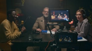 Grabación de un podcast en estudio, con micrófonos profesionales y cámaras enfocando a varias personas sentadas alrededor de una mesa, mientras se transmiten imágenes en una pantalla de la cámara principal.